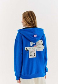 Jasnoniebieska bluza z kapturem z mieszanymi wzorami na plecach, zawierającymi tekst i ilustracje, wykonana z miękkiego materiału o luźnym kroju.