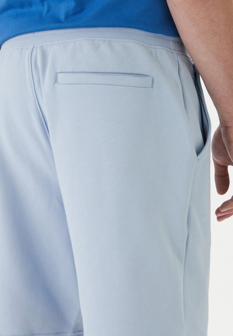 Homme portant un short bleu clair avec une poche passepoilée à l'arrière et une chemise bleu vif rentrée à l'intérieur.