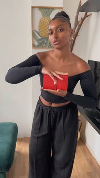 Crvena kožna novčanik s srebrnom kopčom, drži se u rukama. Model nosi crni top bez rukava i crne široke hlače. Pozadina je drveni pod.