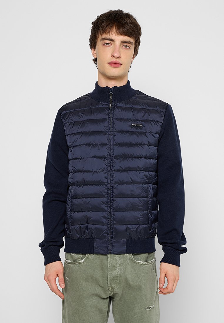 Junger Mann, der eine marineblaue gesteppte Jacke mit gestrickten Ärmeln und Kragen sowie lichtgrünen, abgenutzten Hosen trägt, vor einem schlichten Hintergrund steht.