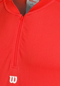 Sudadera deportiva roja con media cremallera. Confeccionada en una tela suave, presenta una textura ligera y un logo blanco de Wilson en el pecho.