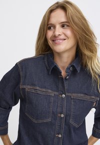 Denimová košile v tmavě modré barvě s oranžovým prošíváním, se límcem, dvěma náprsními kapsami a knoflíkovým zapínáním na přední straně.
