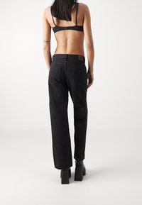 Mujer vestida con jeans rectos de tiro alto negros, sujetador negro y botas de plataforma negras, de pie contra un fondo blanco liso.