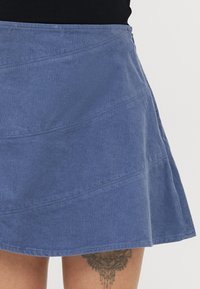 Blå kordfløjlsskjørt med en flagrende design, der har diagonale sømdetaljer og en synlig sidezipper. Glat tekstur og struktureret form.