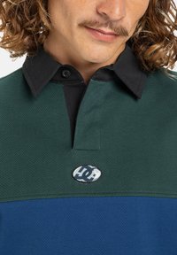 Grøn og blå mesh polo med sort krave, der har en central slids og logo patch. Tekstureret stof skaber en distinkt visuel appel.