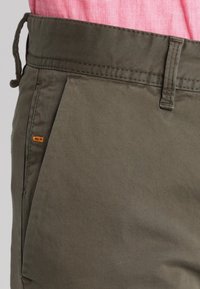 Olivgrüne Hose mit glatter Textur, ausgestattet mit einer Vordertasche und einem orangefarbenen Logoakzent. Einfaches Design, aus leichtem Stoff gefertigt.