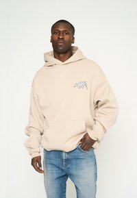 Sweatshirt à capuche beige avec une poche avant, arborant un logo graphique blanc sur la poitrine gauche. Texture douce et coupe décontractée.
