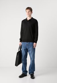 Schwarzes Langarm-Polohemd mit Kragen, kombiniert mit blauen Jeans und schwarzen Lederschuhen. Hält eine schwarze Tragetasche.