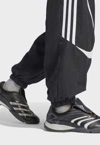 Crne sportske cipele s sivim naglascima, elegantnog dizajna, s gumenim potplatom i elastičnim manšetama oko gležnja, nošene uz crne sužene sportske hlače.