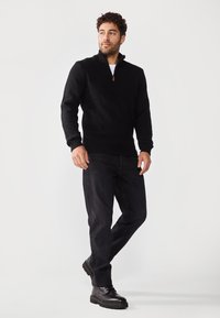 Suéter negro con cuello de cremallera hecho de tejido texturizado, combinado con jeans oscuros y botas negras de cordones. Diseño simple y funcional.