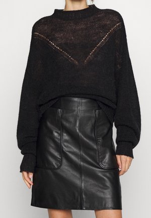 Femme portant un pull en maille noir transparent à manches longues et une jupe en cuir noire taille haute avec poches avant.