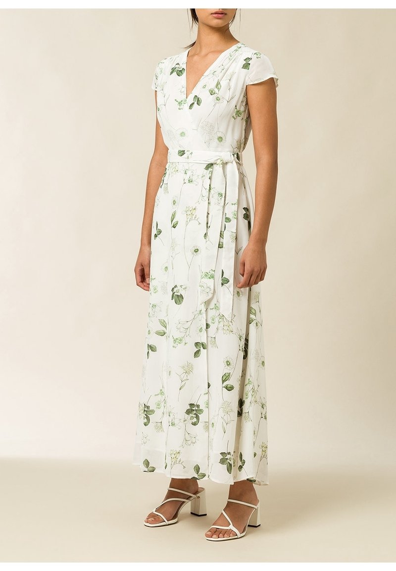 Robe maxi en tissu blanc, ornée de motifs floraux verts. Présente des manches courtes, un design enroulé et un lien à la taille. Sandales à talons blocs blanches.