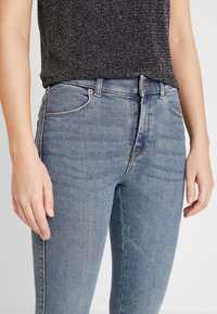 Blå denimjeans med smal passform, hög midja och subtila slitningar. Har klassisk femficksstil och metalldbyxa.