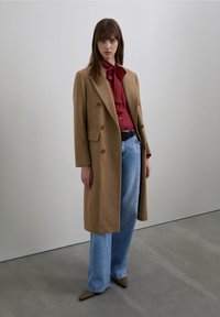 Brown wool double-breasted coat, red blouse with bow tie, light blue wide-leg jeans, and brown pointed-toe shoes against a neutral backdrop.