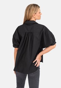 Schwarzes Shirt mit kurzen Ärmeln und Kragen, mit gerafftem Rückendetail und lockerer Passform, aus leichtem Material. Getragen mit dunkelgrauen Jeans.