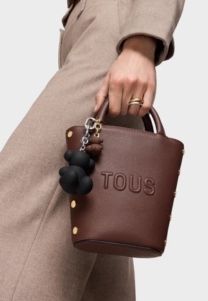 Ręka trzymająca brązową skórzaną torebkę TOUS z złotymi ćwiekami i dwoma brelokami w kształcie misiów, ubraną w beżowy płaszcz oraz nosząca złote i srebrne pierścionki.
