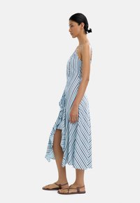 Niebieska i biała w paski sukienka midi z asymetrycznym dołem, cienkimi ramiączkami oraz falbaną z boku. Nosi ją z brązowymi płaskimi sandałami.