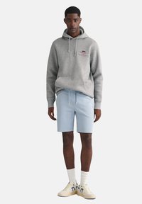 Sweatshirt à capuche gris avec cordons, shorts bleu clair, chaussettes blanches à mi-mollet et baskets beiges avec accents bleu foncé.