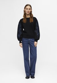 Pull noir avec une broderie florale texturée bleu foncé sur les manches bouffantes, associé à un jean large bleu et des chaussures noires.