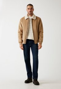 Beige Bomberjacke mit einem Kunstfellkragen, getragen über einem strukturierten cremefarbenen Pullover. Dunkelblaue gerade Jeans und dunkle Schuhe runden den Look ab.