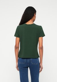 Σκούρ�ο πράσινο κοντομάνικο μπλουζάκι με στρογγυλή λαιμόκοψη, ελαφρώς χαλαρή εφαρμογή και ραμμένο στρίφωμα, συνδυασμένο με μπλε τζιν.