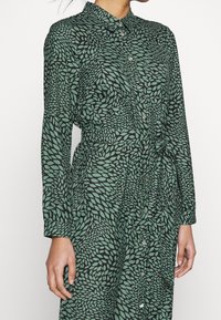 Femme portant une robe verte à manches longues avec des motifs de feuilles noires, fermeture par boutons à l'avant, col et ceinture en tissu nouée à la taille.