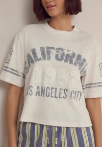 Bílý zkrácený tričko s modrým opotřebovaným textem "CALIFORNIA 1982 LOS ANGELES CITY" a pruhy na rukávech; měkký bavlněný materiál.