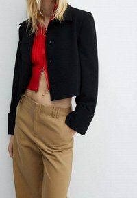Zwarte cropped jas met lange mouwen, gecombineerd met een rode ribbeltop en beige broeken met een gladde textuur en een getailleerd ontwerp.