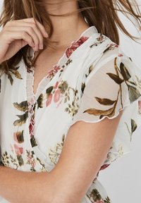 Kukkakuosiset puserot, joissa on valkoista läpikuultavaa kangasta, sisältävät vaaleanpunaisia ja vihreitä kukkakuvioita, röyhelösateet ja herkän kaula-aukon.