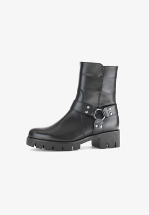 Stivale alla caviglia in pelle nera con suola spessa, tacco a blocco e cinturino decorativo con anello metallico e ribattini attorno alla caviglia.