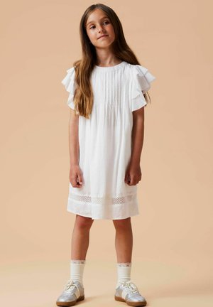 Fetiță cu păr lung, purtând o rochie albă cu mâneci cu volane, șosete din dantelă și teniși argintii, stând pe un fundal bej.