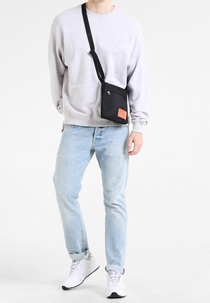 Sort crossbody-taske med lynlåslomme foran og orange logopatch, båret over en grå sweatshirt og lyseblå slidte jeans.