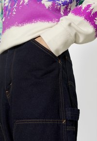 Pantalones vaqueros de tono azul oscuro, con costuras visibles, un bolsillo lateral y una mano descansando en el bolsillo delantero.