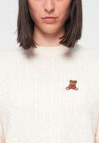 Pulover tricotat de culoare crem, cu un model textural de cablu, având un mic broderie cu un ursuleț de pluș maro pe pieptul stâng.