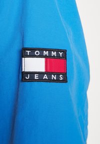 Blue fabric with a square patch displaying "TOMMY" in white text, a red and white rectangular stripe, and "JEANS" in white on a black background.