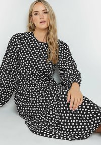 Vestido negro con lunares blancos, mangas largas y abullonadas, cintura fruncida y falda fluida. Tela suave con una textura lisa.