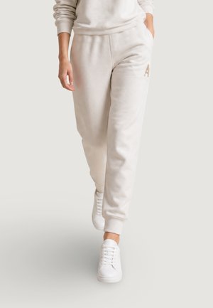 Personne portant un pantalon de jogging beige clair et des baskets blanches marchant, montrée de la taille aux pieds sur un fond uni.