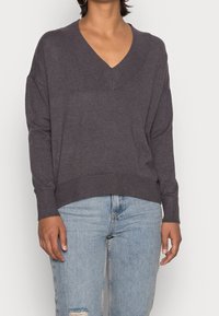 Grauer, weicher Strickpullover mit tiefem V-Ausschnitt, überschnittenen Schultern sowie gerippten Bündchen und Saum. Kombiniert mit hellblauen, verwaschenen Jeans.