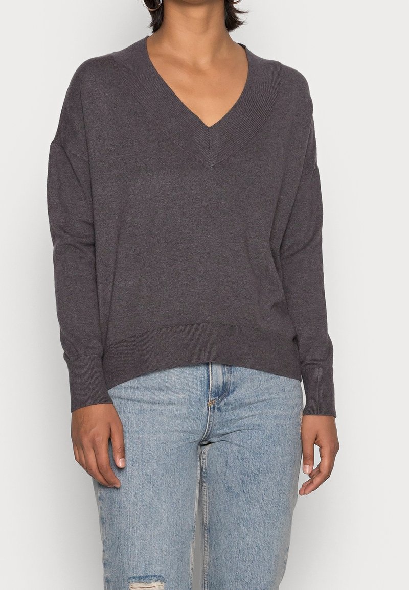 Grauer, weicher Strickpullover mit tiefem V-Ausschnitt, überschnittenen Schultern sowie gerippten Bündchen und Saum. Kombiniert mit hellblauen, verwaschenen Jeans.