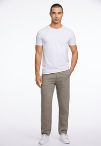 Camiseta blanca de manga corta y cuello redondo combinada con pantalones de textura beige claro, modelo de pie con las manos en los bolsillos y zapatillas blancas.