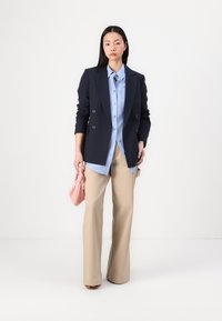 Blazer blu navy doppiopetto sopra una camicia azzurro chiaro con bottoni e pantaloni beige a gamba larga, mentre tiene in mano una piccola borsa rosa.