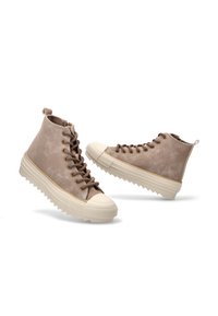 Zapatillas beige tipo high-top con gruesas suelas de goma blanca y punteras, con cordones marrones y una suela texturizada para mayor agarre.