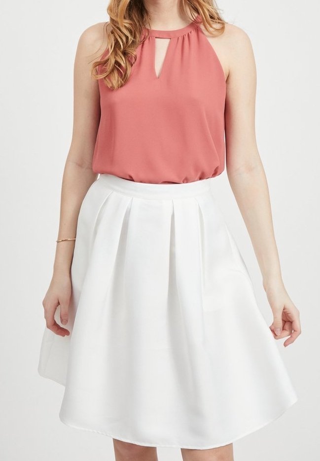 Femme portant un haut rose sans manches avec un décolleté en forme de trou de serrure et une jupe blanche plissée taille haute, debout devant un fond uni.