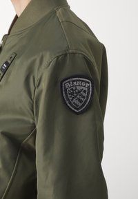 Olivově zelená bomber bunda s žebrovaným límcem a ramenním znakem, který má složité korálkování ve tvaru štítu. Hladká, nylonová textura.