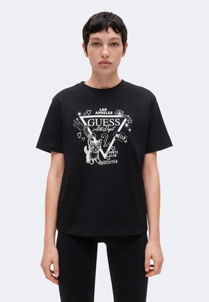 Personne portant un T-shirt noir Guess avec des motifs blancs comprenant un chaton, des cœurs, des étoiles et un texte faisant référence à Los Angeles et à la Californie.
