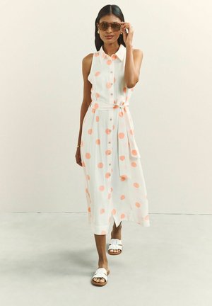 Nő fehér ujjatlan ruhát viselve, narancssárga pöttyökkel, fehér szandálban és barna napszemüvegben előrefelé sétál, egyszínű bézs háttér előtt.