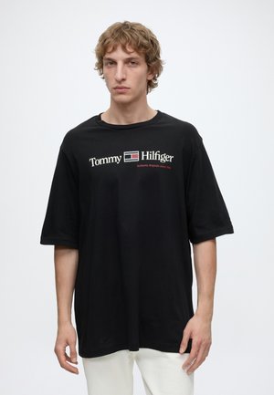 Jeune homme aux cheveux clairs et bouclés porte un t-shirt noir ample Tommy Hilfiger avec un logo blanc et rouge, debout devant un fond blanc.