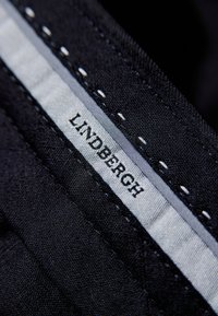 Zwarte stoflabel met het merk "LINDBERGH" in witte letters, voorzien van een contrasterende grijze streep en witte stiksels.