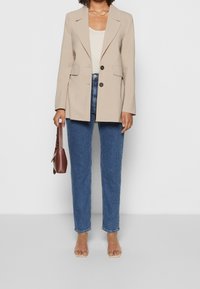 Beige oversized blazer with two black buttons, paired with light blue high-waisted jeans and transparent heeled sandals. Holding a brown handbag.
