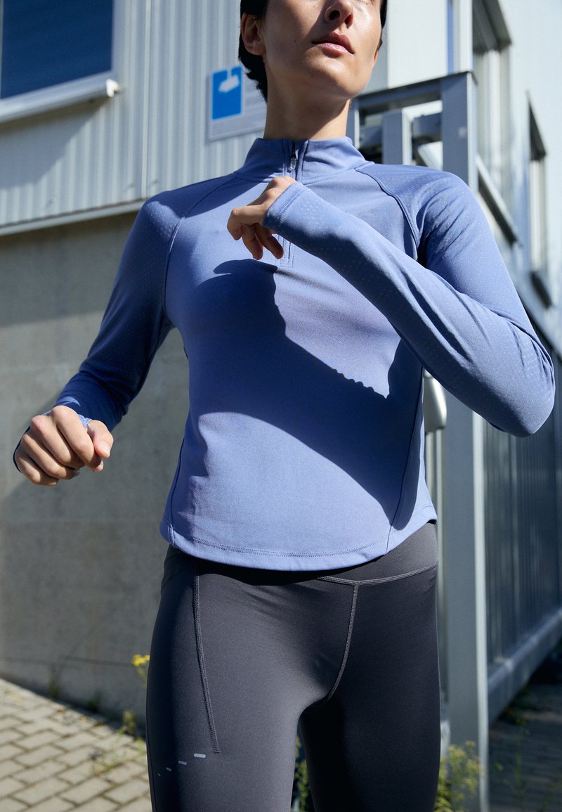 Lyseblå langermet treningsgenser med delvis glidelås, kombinert med tettsittende mørkegrå leggings med horisontale linjedetaljer.
